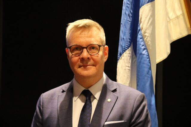 Kuvassa etualalla Tuomas Kuusivaara puvussa. Kuvan taka-alalla tummaa taustaa vasten on Suomen lippu.